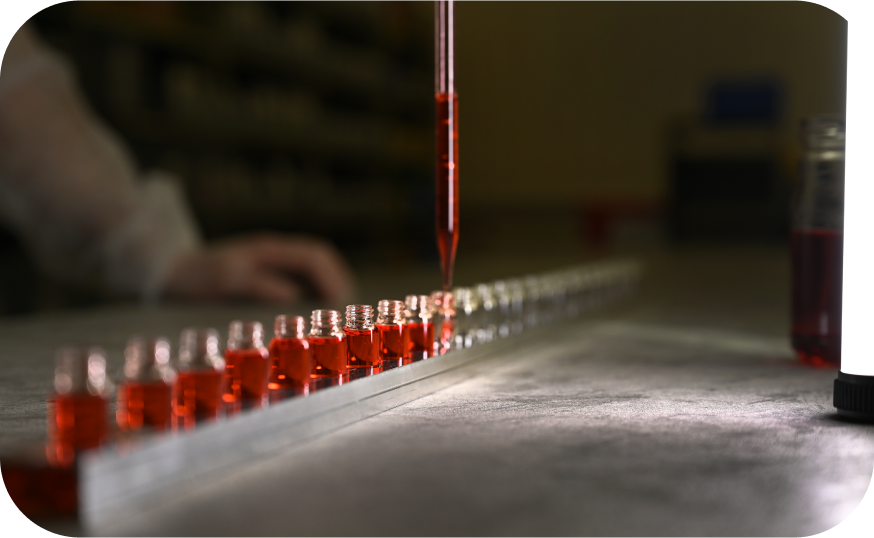 Mehrere kleine Glasfläschchen, gefüllt mit roter Flüssigkeit, stehen in einer Reihe auf einem metallischen Gestell. Eine Pipette tropft rote Flüssigkeit in eines der Fläschchen. Im Hintergrund ist eine Hand in einem weißen Laborkittel unscharf zu sehen. Am rechten Bildrand befindet sich eine Lichtquelle und ein weiteres Fläschchen mit roter Flüssigkeit. Die Szene spielt offenbar in einem Laborumfeld.