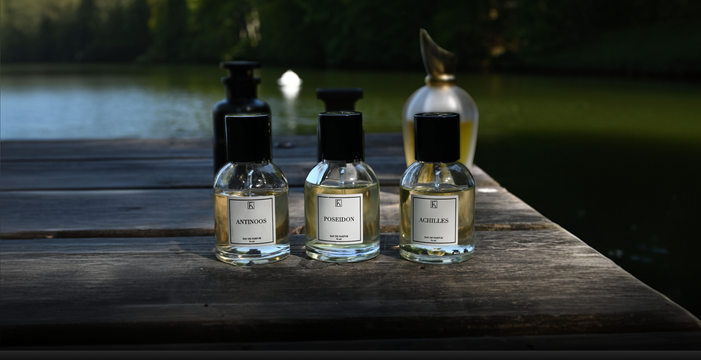 Auf einem hölzernen Steg stehen fünf Parfümflaschen, drei vorne mit den Aufschriften „Antinoos“, „Poseidon“ und „Achilles“, zwei weitere, größere Flakons weiter hinten. Der Steg befindet sich an einem ruhigen See, im Hintergrund sind Bäume und Wasser erkennbar, mit einem unscharfen weißen Objekt im See. Die Szenerie ist natürlich, das Licht ist sanft und vermittelt eine entspannte Atmosphäre am Wasser.