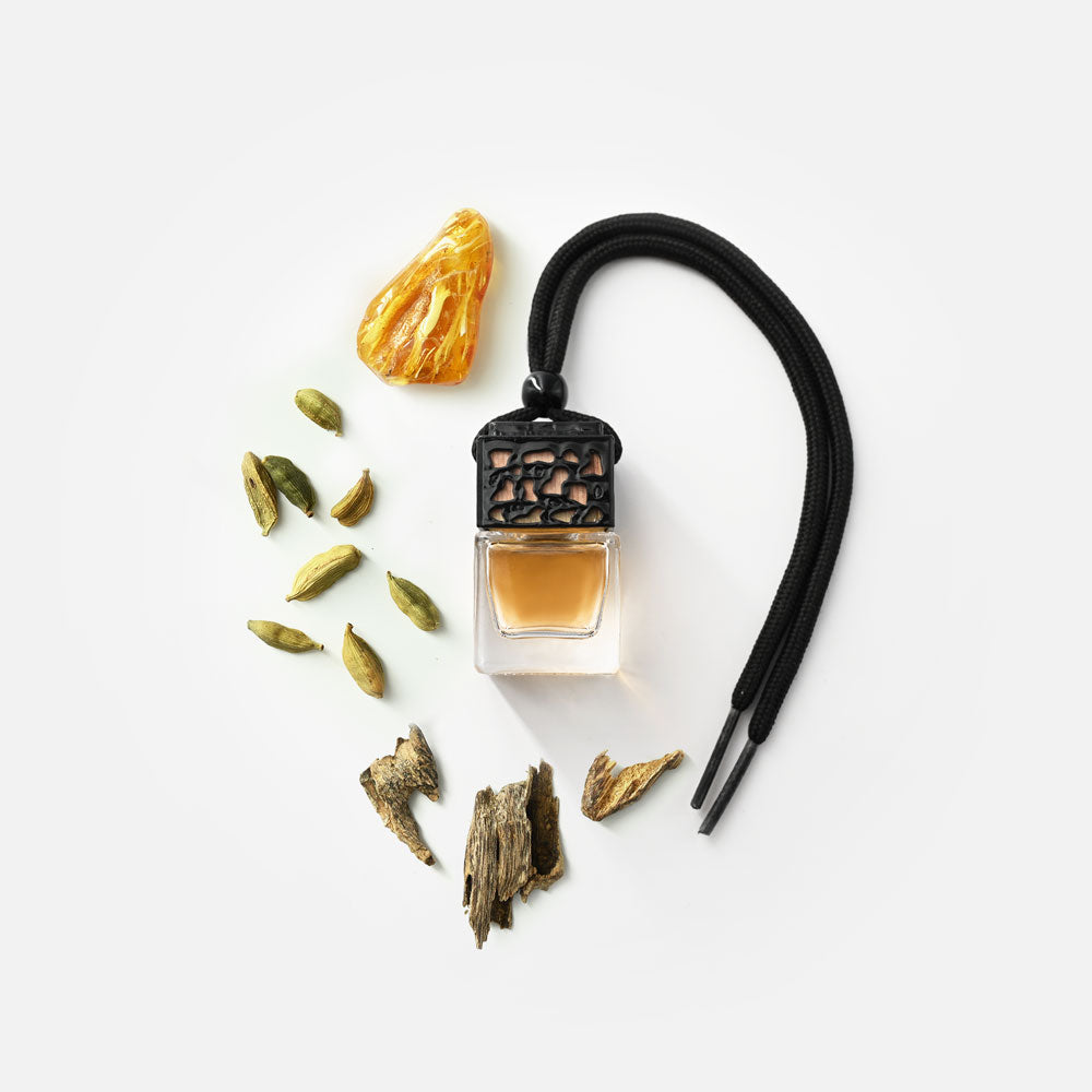 Ein kleines rechteckiges Glasfläschchen mit schwarzem Verschluss und einer schwarzen Kordel, umgeben von getrockneten grünen Kardamomkapseln, braunen Holzstücken und einem großen gelb-orangen Harzstück, alles auf weißem Hintergrund arrangiert.
