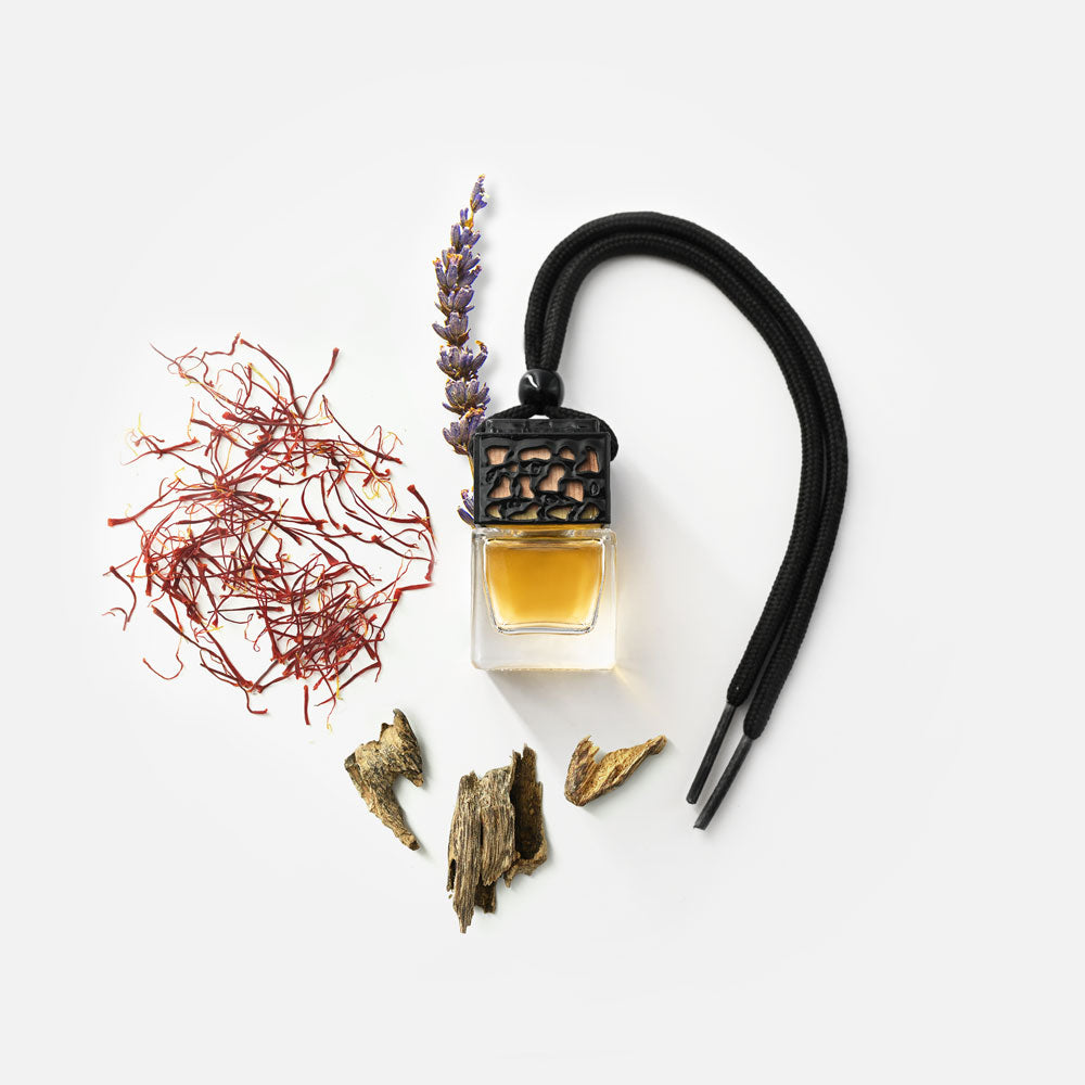 Ein kleines Fläschchen Autoduft mit schwarzem Deckel und Band, daneben Lavendelblüte, Holzstücke und rote Safranfäden.